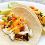 Close up of tacos filled with grilled halibut filet and peach salsa with lime wedges on the side.