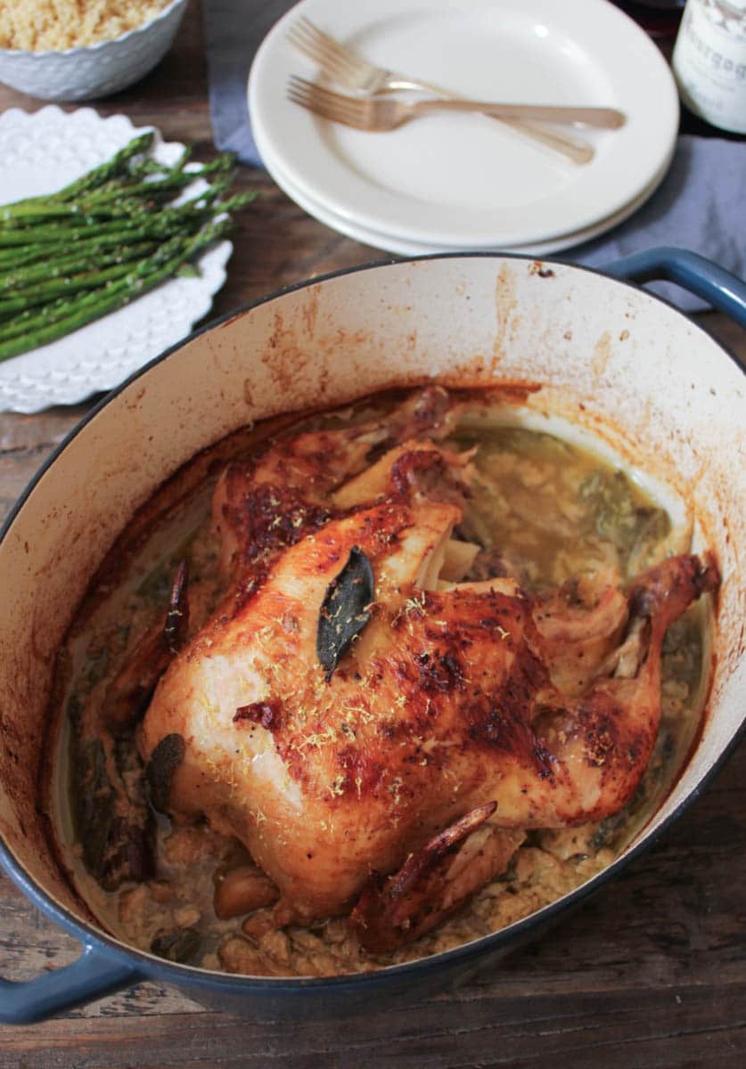 Overhead view of a whole roasted chicken in milk with quinoa and asparagus on plates in the background.
