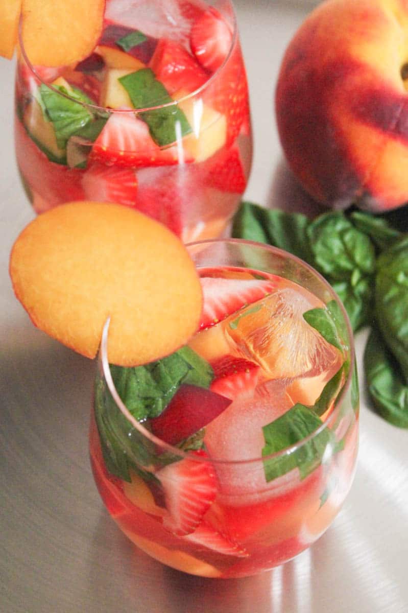Overhead view of two glasses of rose sangria with floating strawberries, peaches and basil on a tray next to a whole peach.