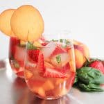 Two glasses of sparkling rose sangria garnished with thin peach slices in front of a display of summer fruits.