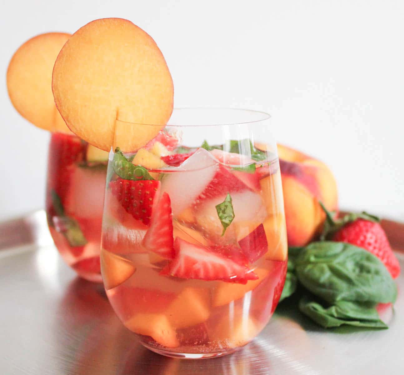 Two stemless wine glasses filled with sparkling rose sangria containing strawberries, peaches, and basil on a table displaying the fruits in their whole forms. 