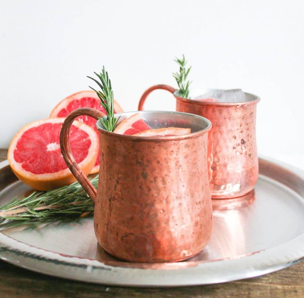 Two cups of Grapefruit Rosemary Mule in moscow mule cups on a platter with grapefruit halves and rosemary sprigs. 