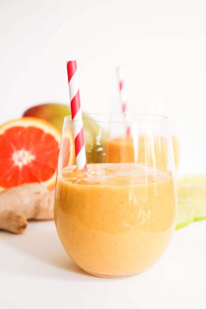 Glass of orange smoothie with red and white spiral straw behind are ginger root , a mango, and a halved orange.