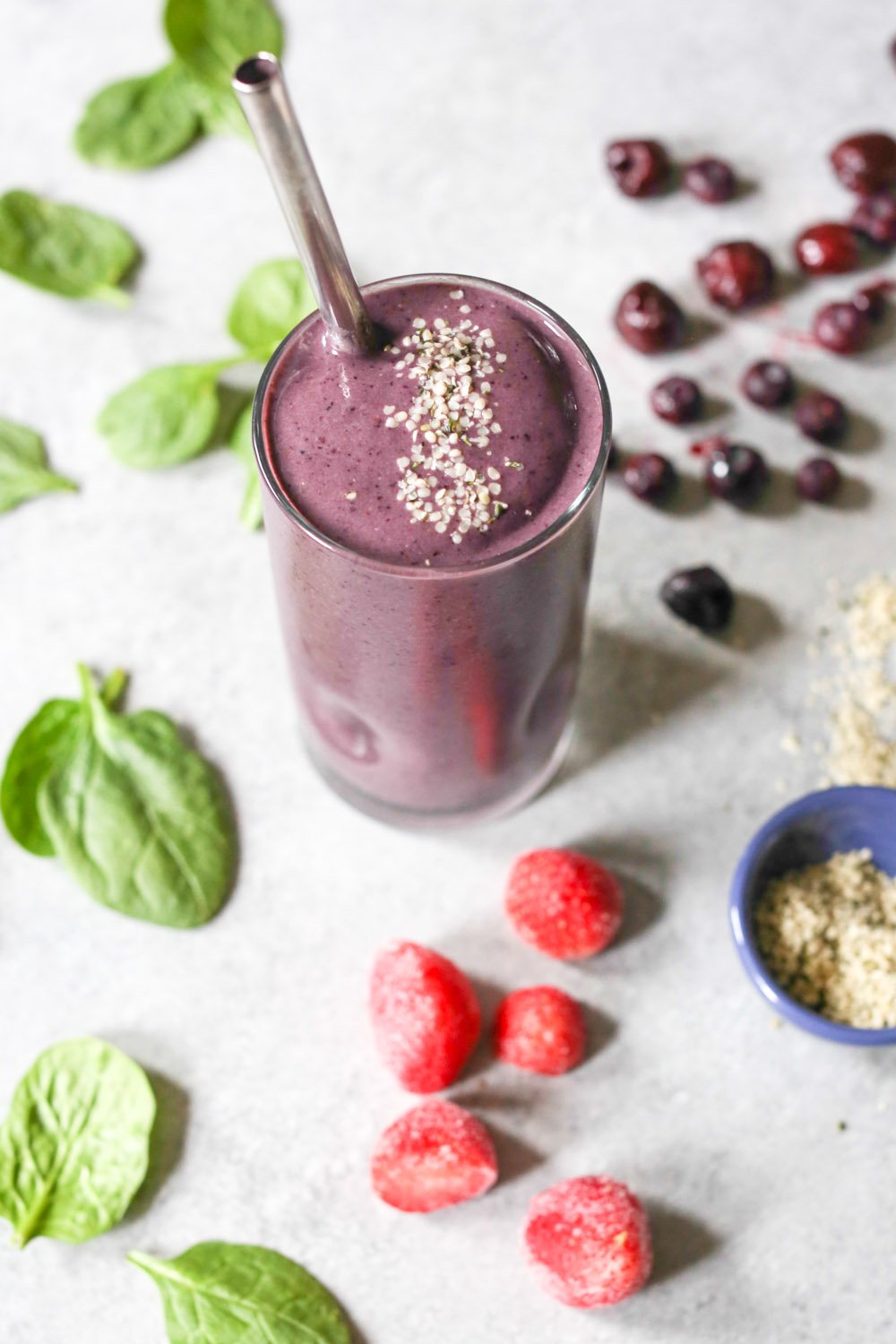 Purple berry cherry smoothie in a glass with a metal straw surrounded by spinach leaves, berries, and bowl of seeds.