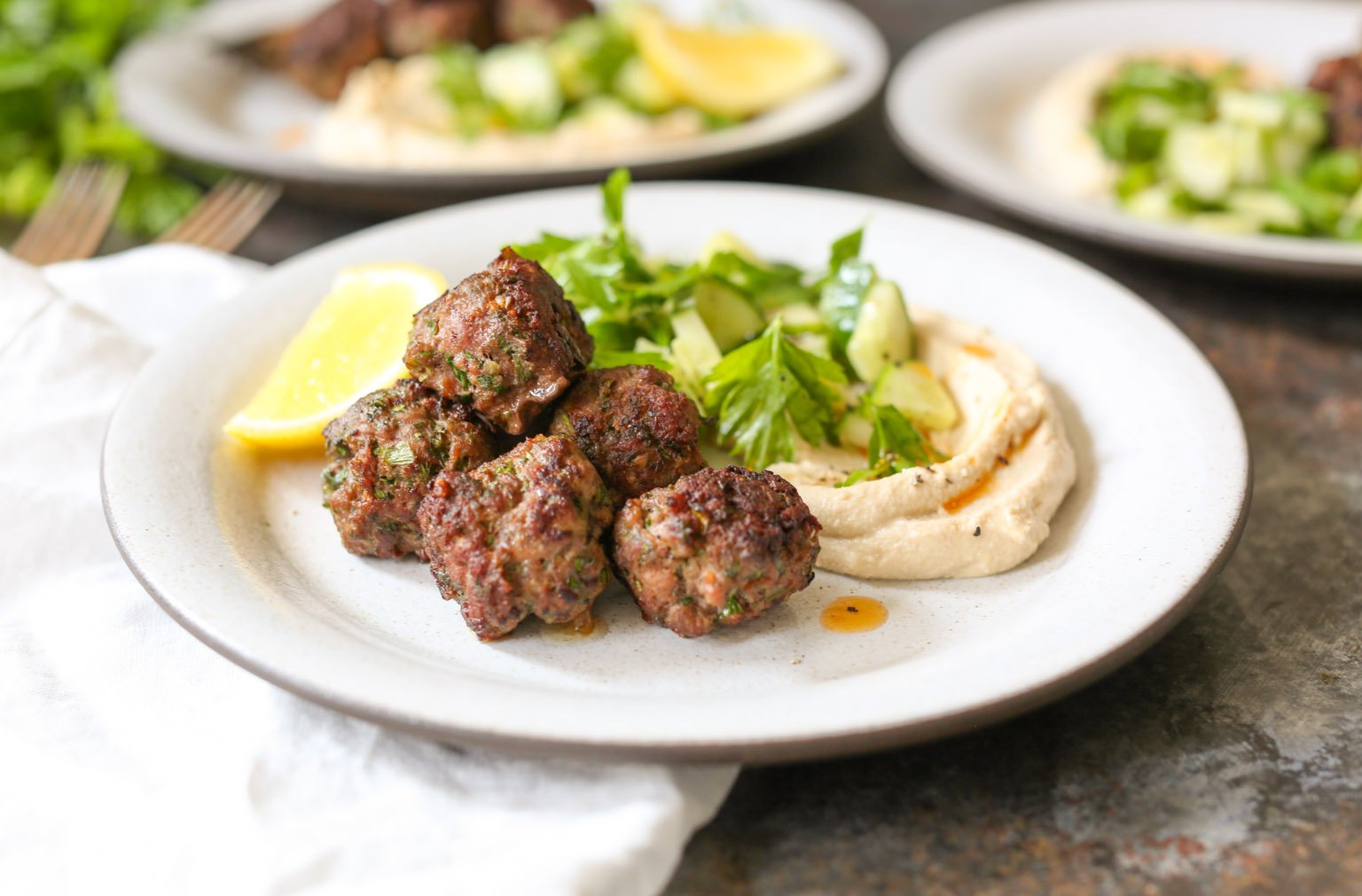 Side view of a plate of spiced lamb meatballs with hummus and a lemon wedge.