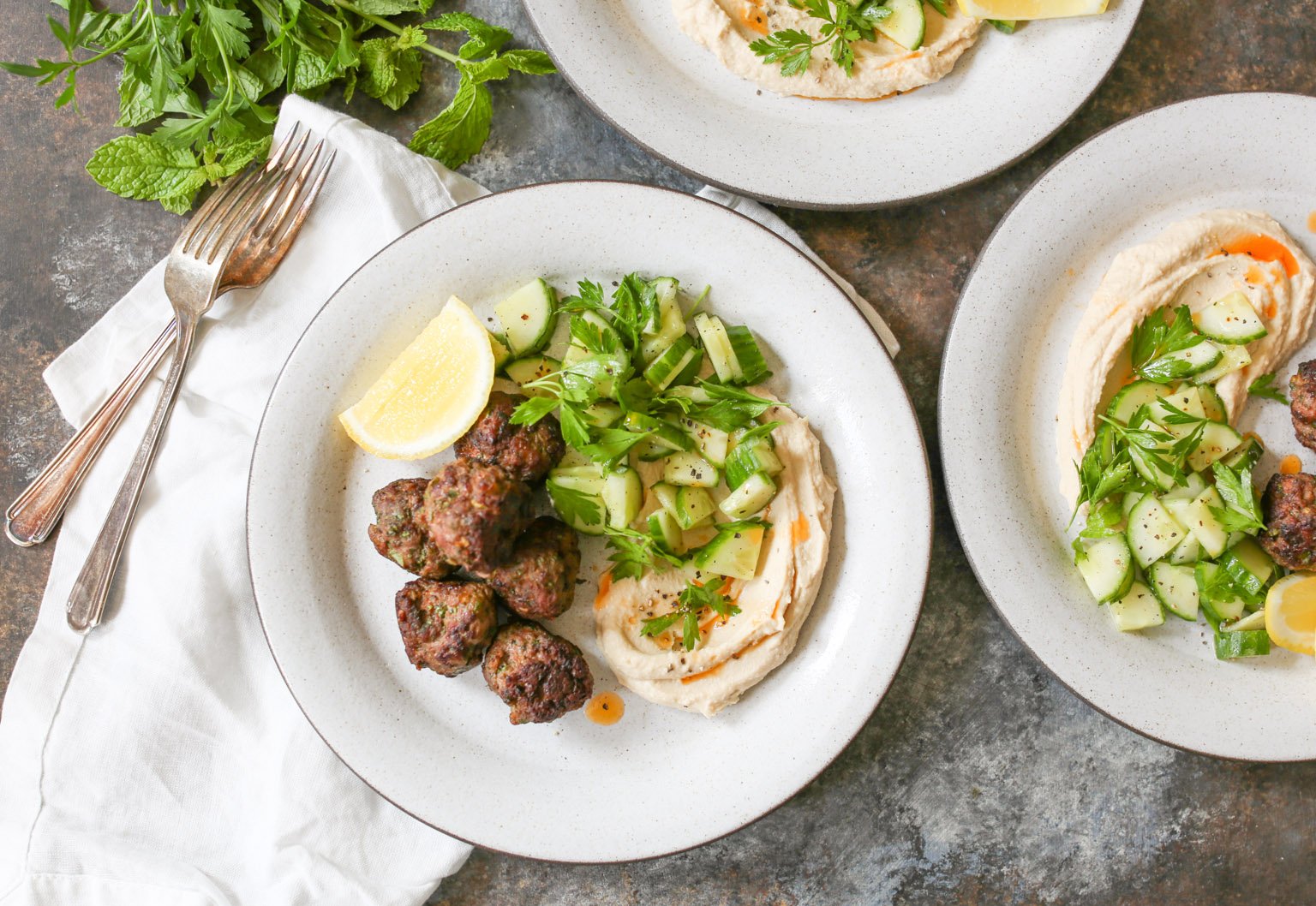 Overhead view of plated lamb kofta with hummus, cucumber salad, and lemon with napkins and forks. 