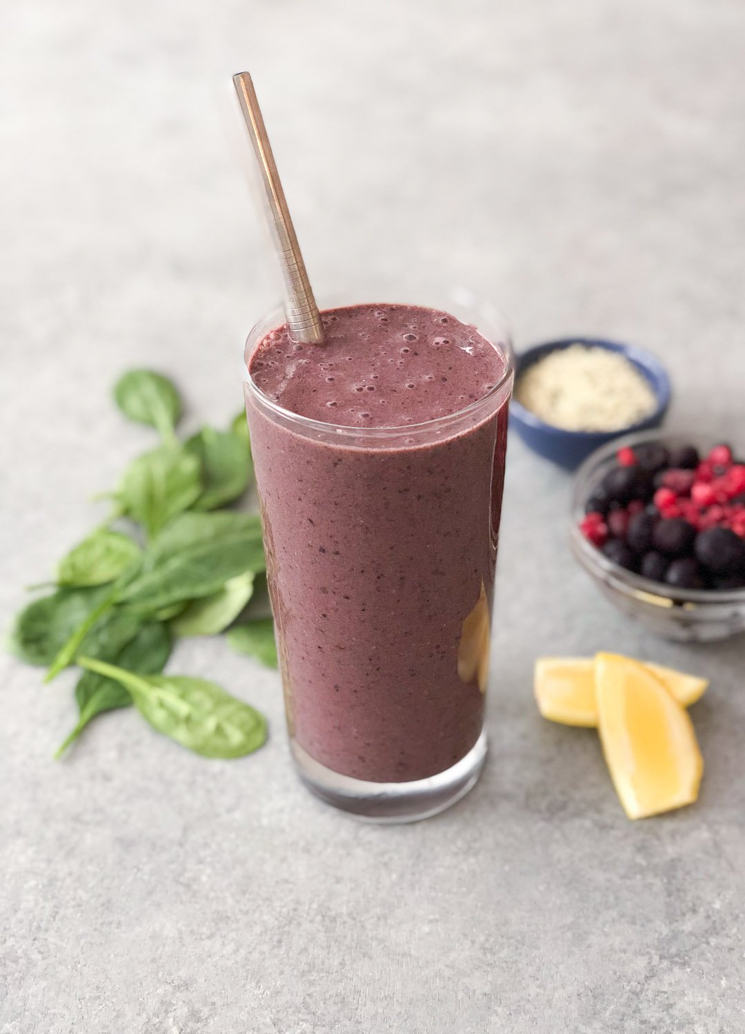 Purple colored smoothie in a glass with a metal straw. The cup is surrounded by spinach leaves , lemon slices, and bowls of fruit and seeds. 