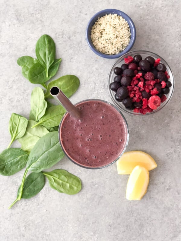 Glass of berry chia seed smoothie with a metal straw surrounded by a dish of seeds, a dish of frozen berries, a couple slices of lemon, and spinach leaves.
