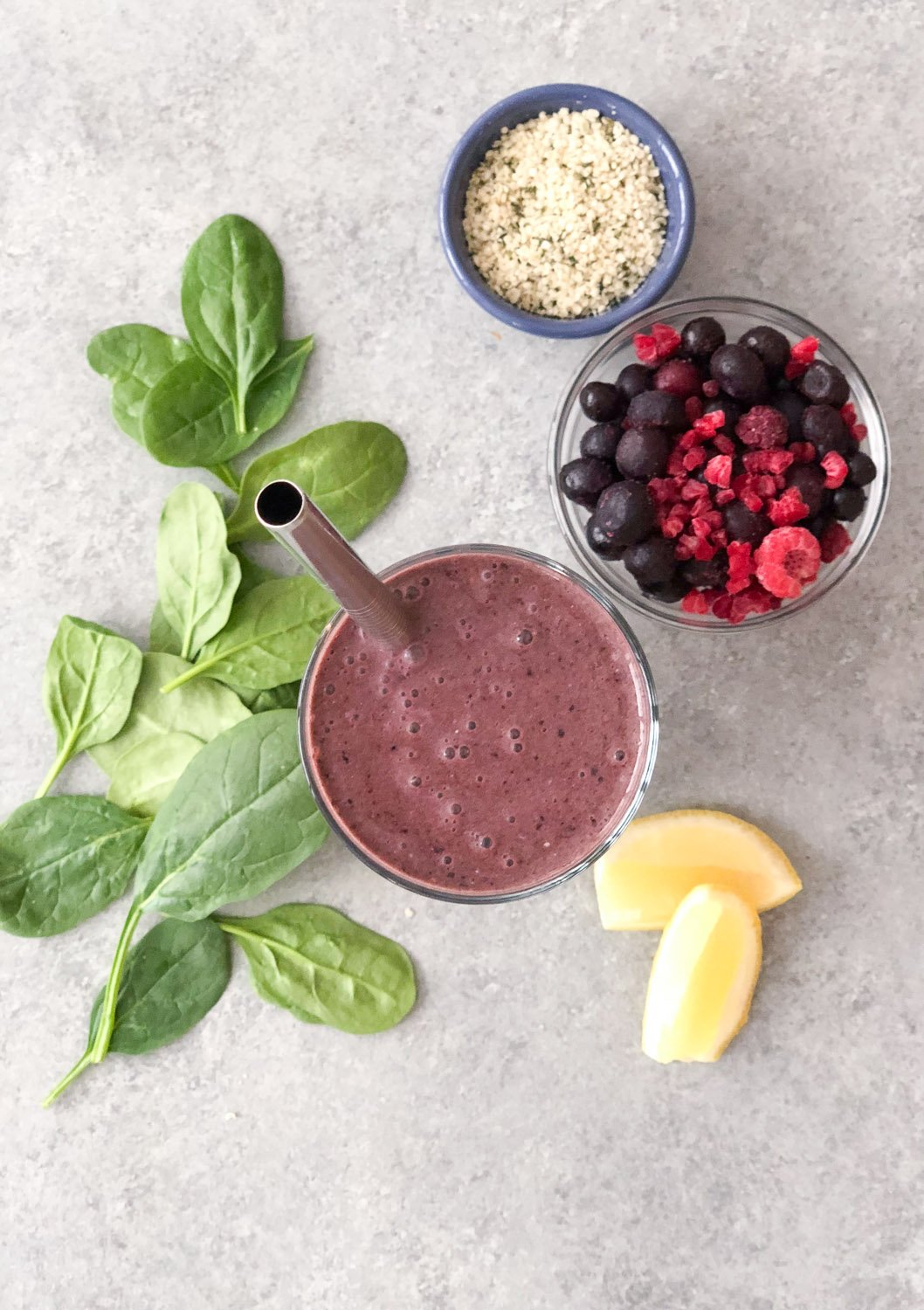 Glass of green berry smoothie from above with metal straw surrounded by a dish of seeds, dish of frozen berries, a couple slices of lemon, and some spinach leaves.