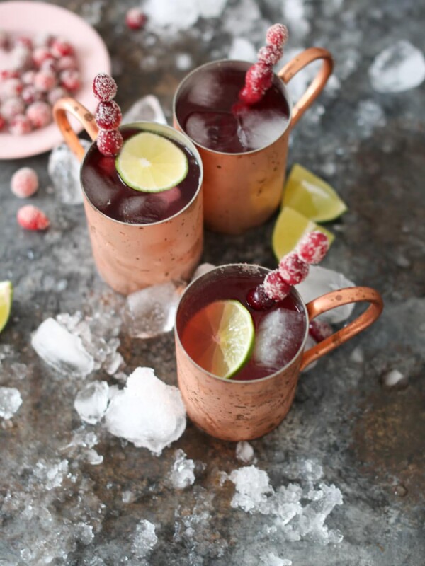 Overhead shot of three cranberry moscow mules in copper mugs with sugared cranberry swizzle sticks and bowl of sugared cranberries.