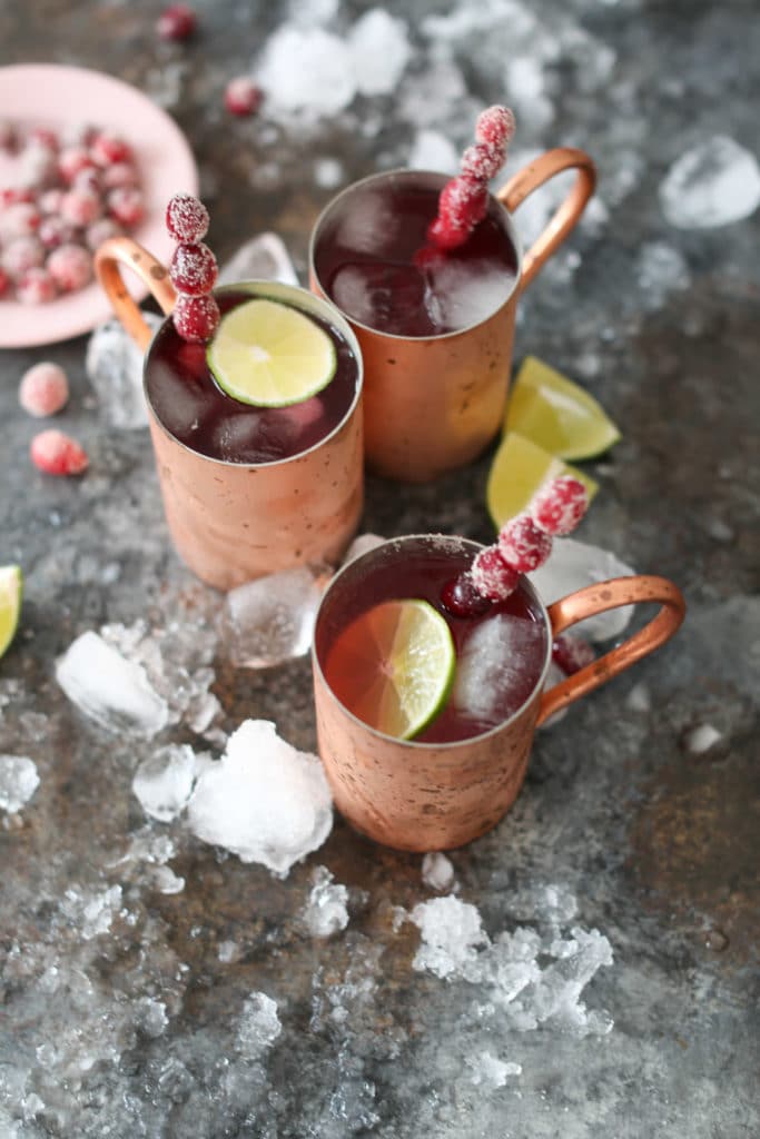 Overhead shot of three cranberry moscow mules in copper mugs with sugared cranberry swizzle sticks and bowl of sugared cranberries.