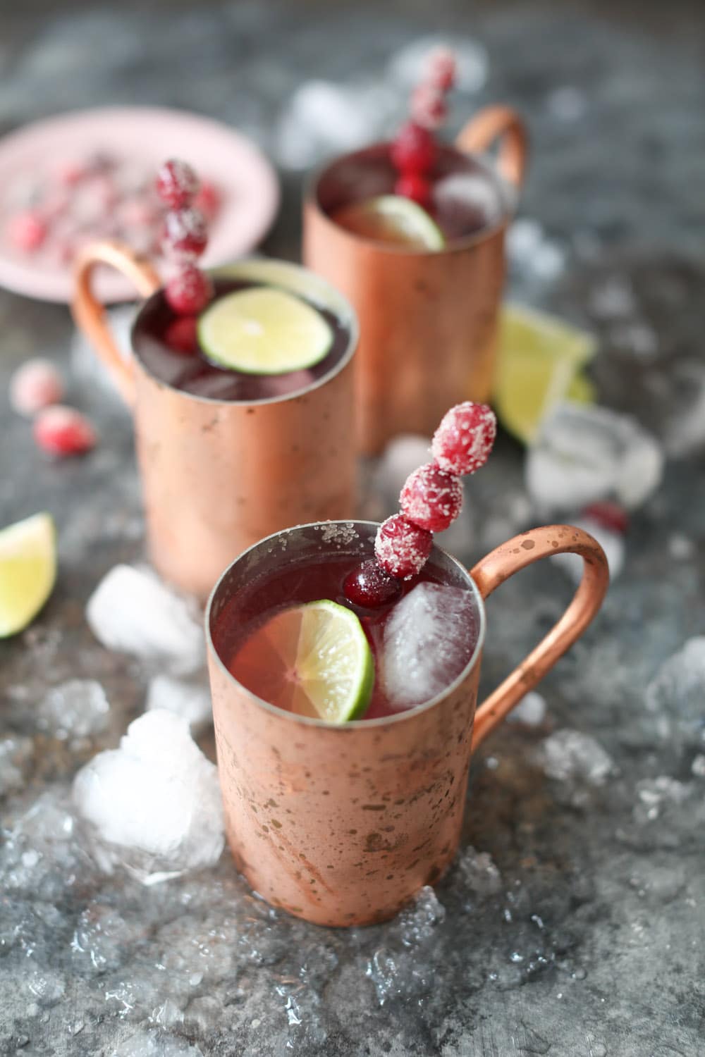 Close-up of a refreshing cranberry Moscow mule in a copper mug with two slightly blurred cocktails in the background. 