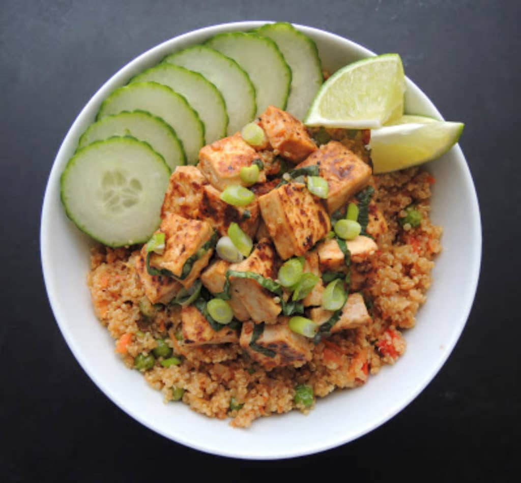 Bowl with basil tofu on a bed of stir fried quinoa garnished with sliced cucumber and lime wedges.
