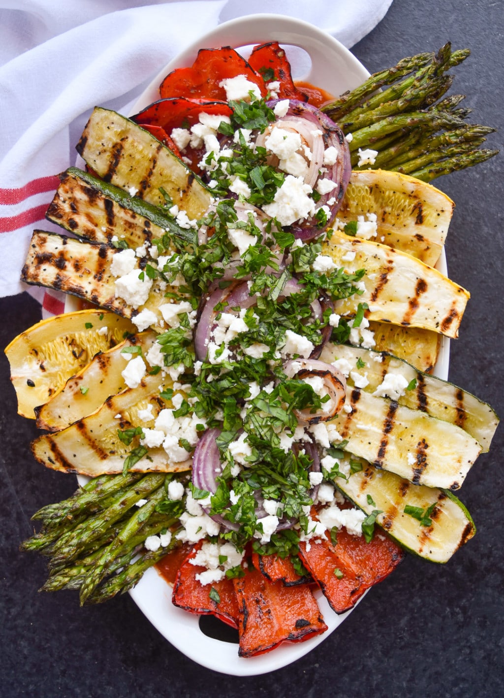 Close up of platter of grilled zucchini, yellow squash, onions, bell pepper, and asparagus topped with feta cheese and chopped herbs.