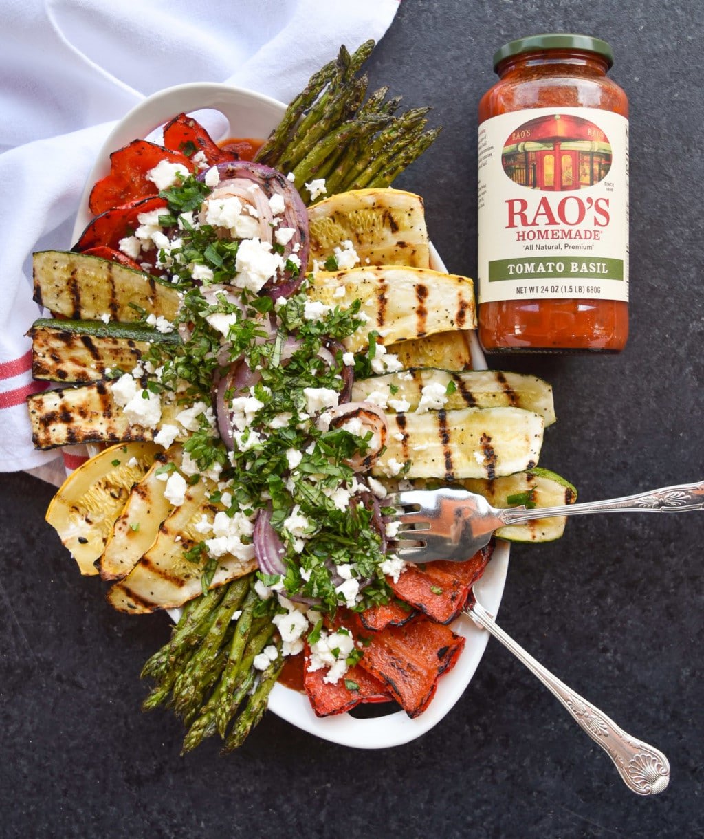 Platter of colorful grilled vegetables with serving utensils next to a full jar of Rao's Tomato Basil Sauce.
