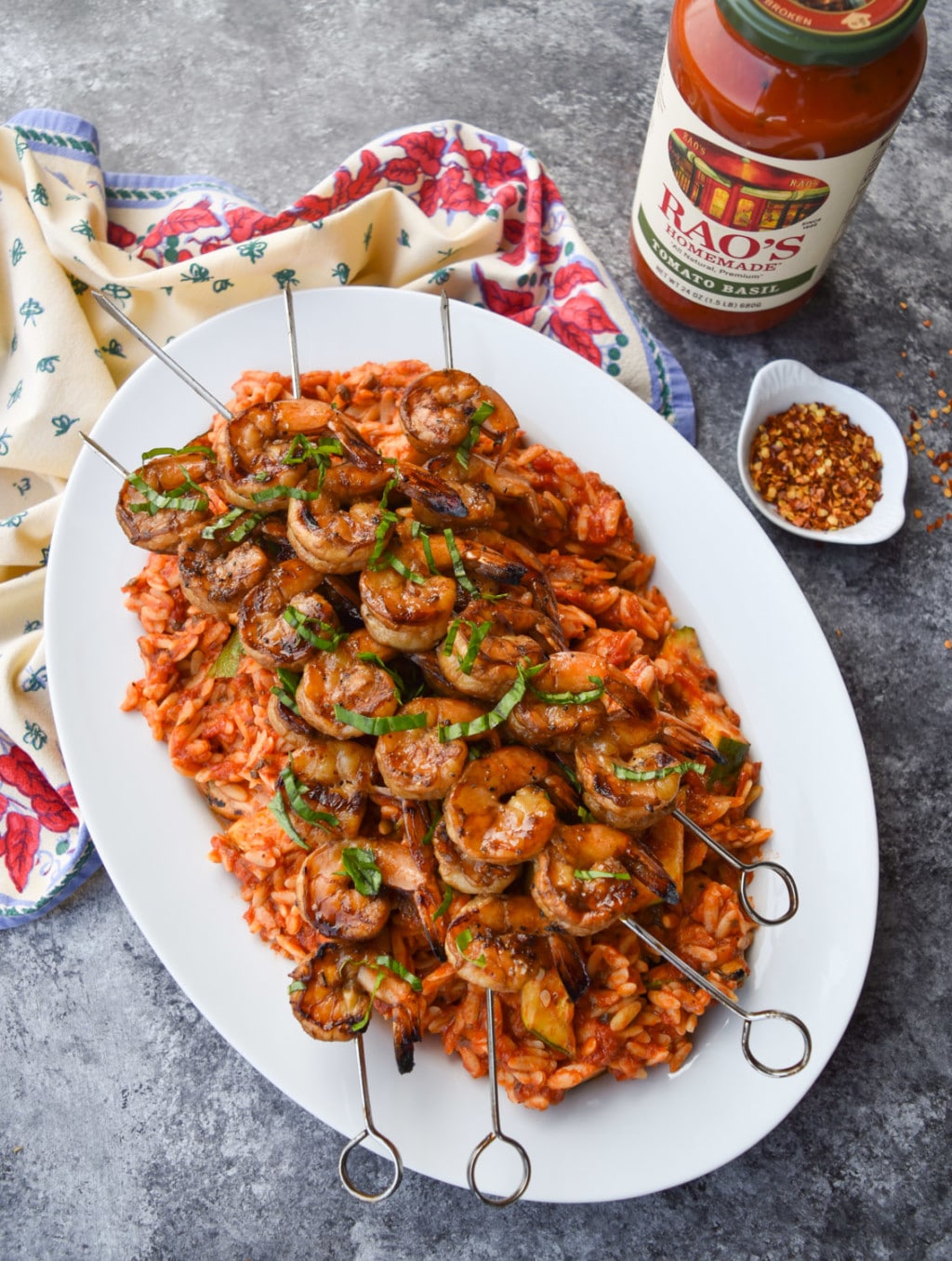 Horizontal platter of orzo with tomato sauce topped with four skewers of grilled shrimp alongside a jar of Rao's Homemade Tomato Basil Sauce.
