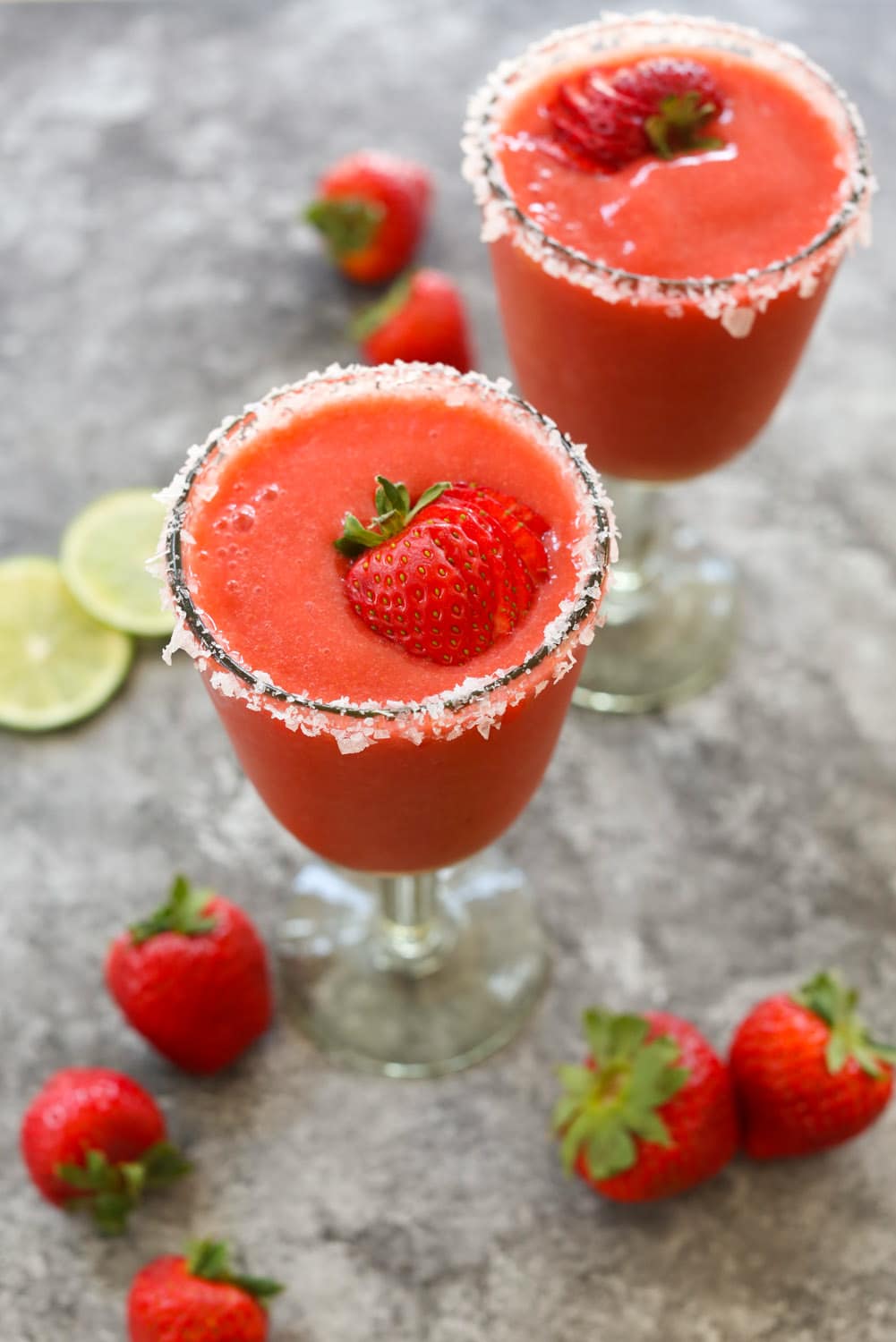 One frozen strawberry margarita with a salted rims in the foreground with a second margarita in the background along with decorative strawberries and lime rounds.
