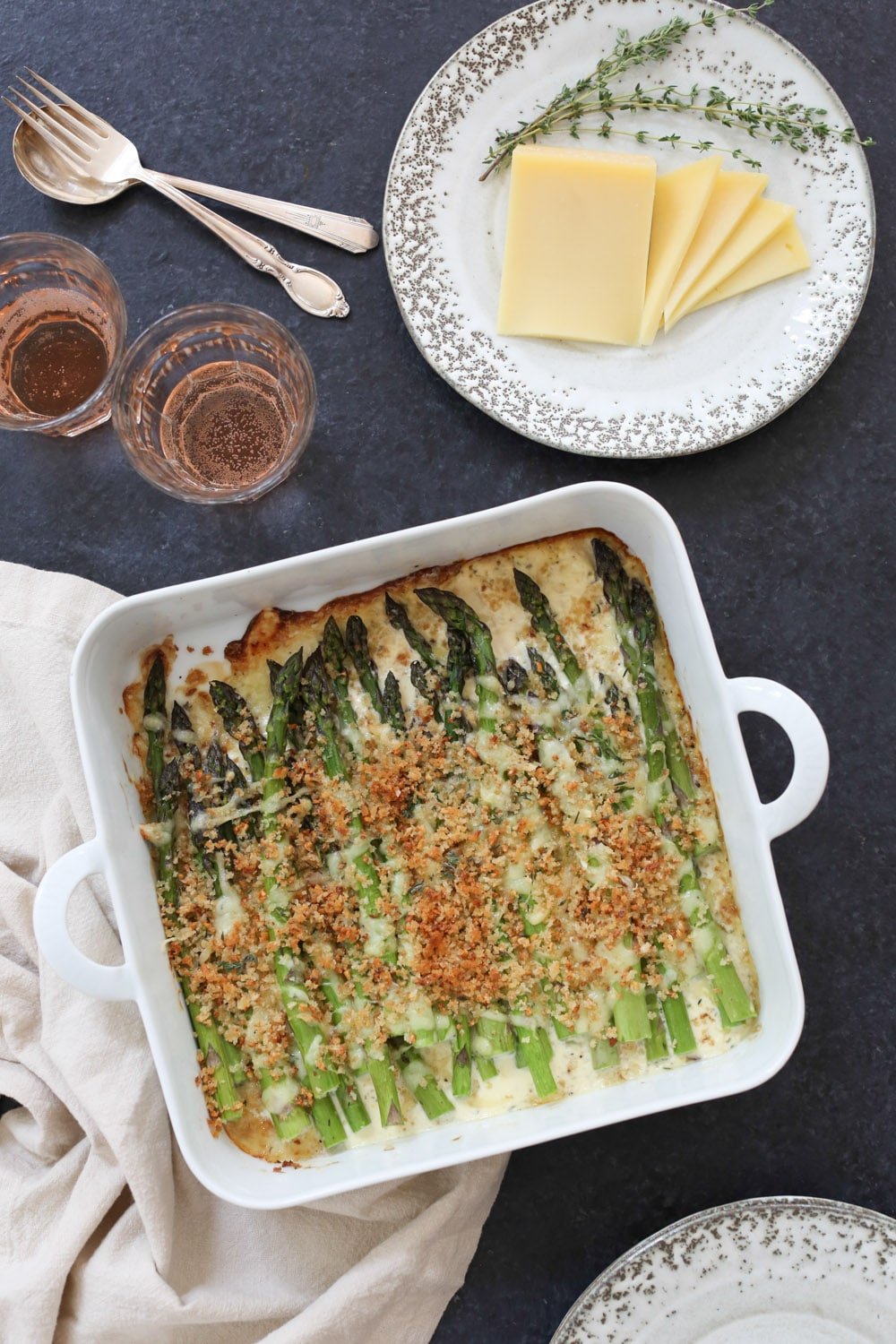 Overhead image of baked asparagus gratin with a plate of sliced Austrian
Mountain cheese and two glasses of sparkling rosé.
