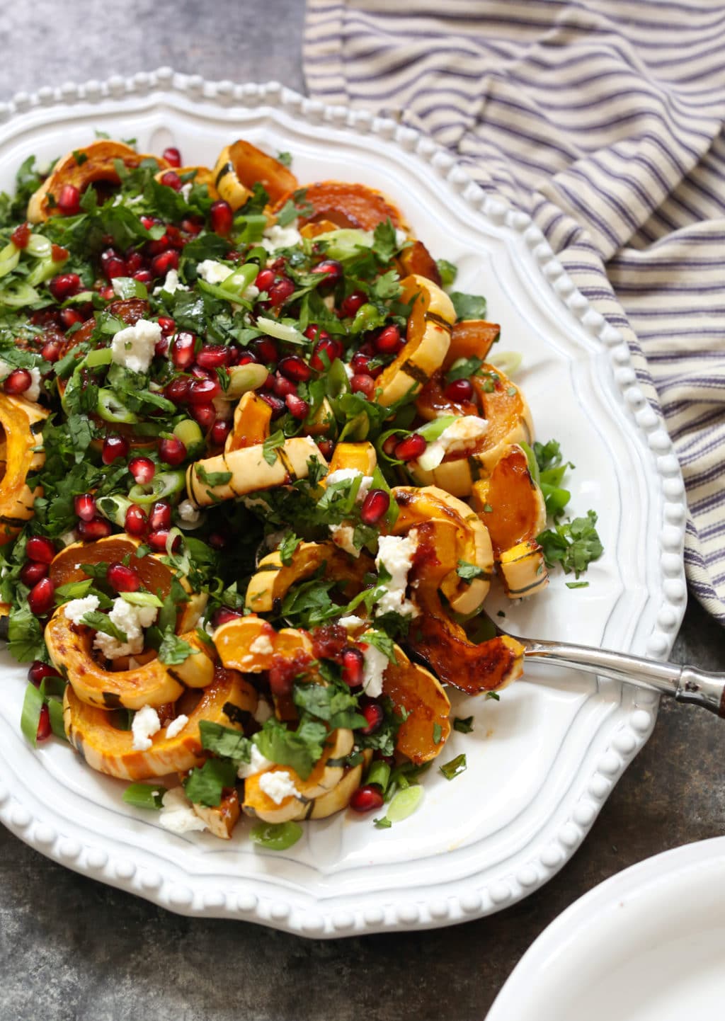Partial side view of roasted delicata squash topped with herbs, goat cheese, and pomegranate on a white serving platter with a spoon scooping out a single portion.