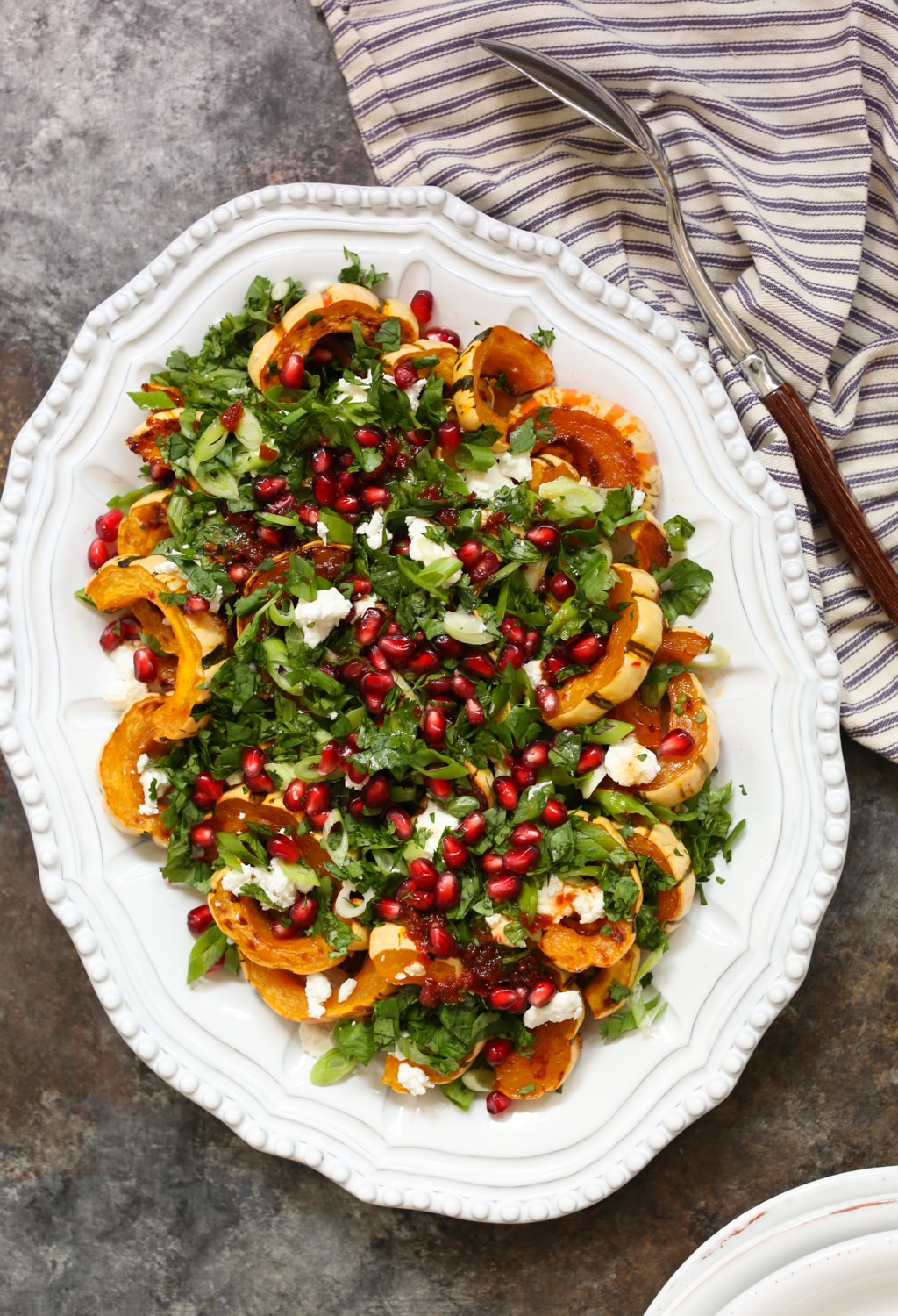 Overhead view of roasted chipotle delicata squash with herbs, goat cheese, and pomegranate on a platter with a serving spoon off to the side.