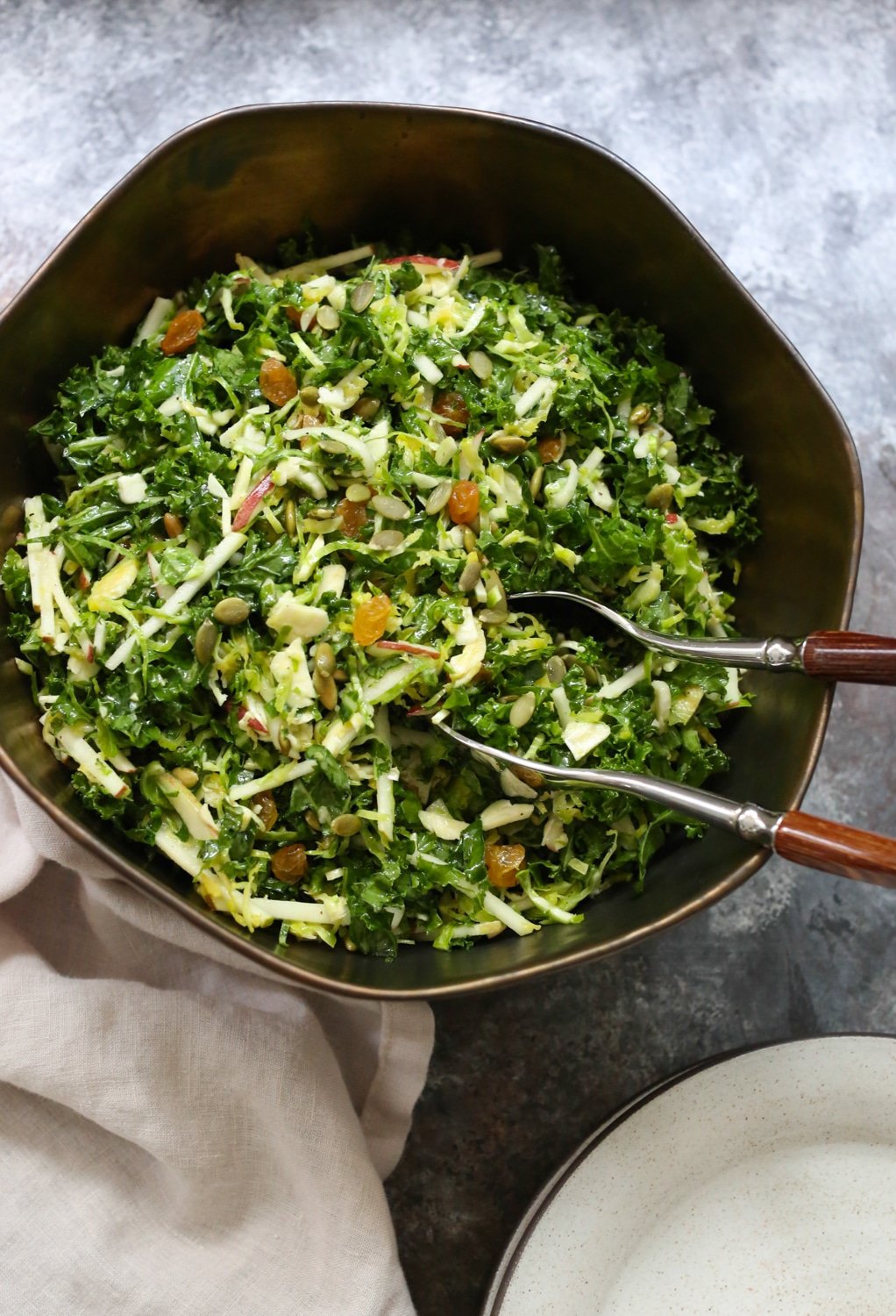 Overhead closeup of Kale, Brussels Sprout, and Apple Salad in a bowl with serving utensils in the bowl and side plates in the bottom right corner. 