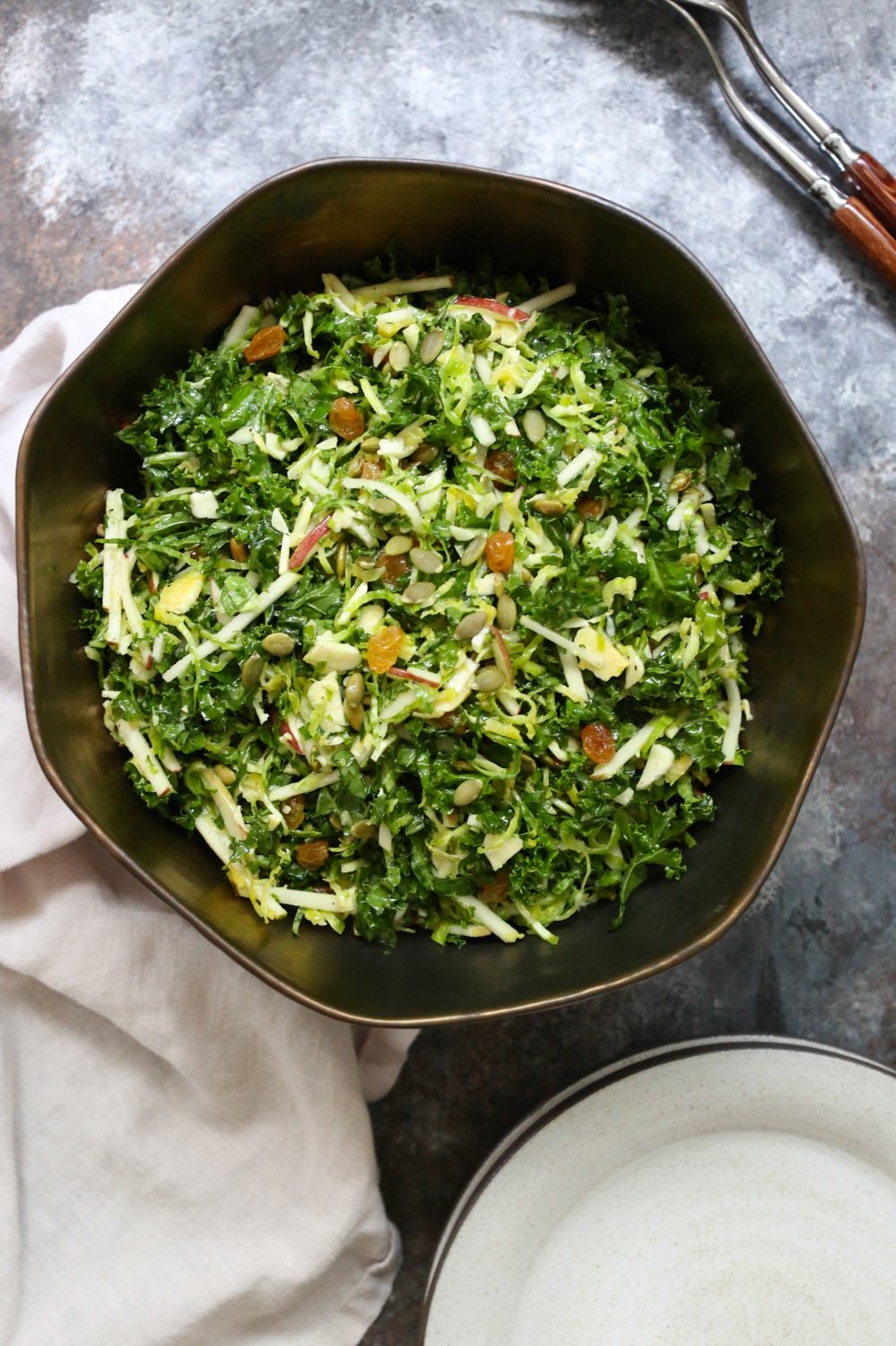 Overhead view of Kale, Apple, and Brussels Sprouts Salad in a large bowl with some serving utensils peeking into the upper right corner. 